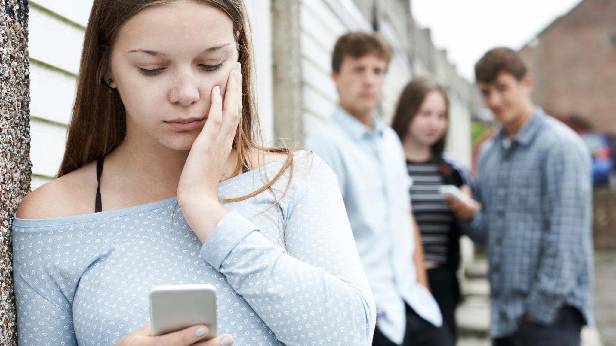 Junge Frau steht absteits einer Gruppe mit drei jungen Menschen und blickt auf ihr Handy. Es wirkt, als ob die Gruppe abfällig über die spricht.