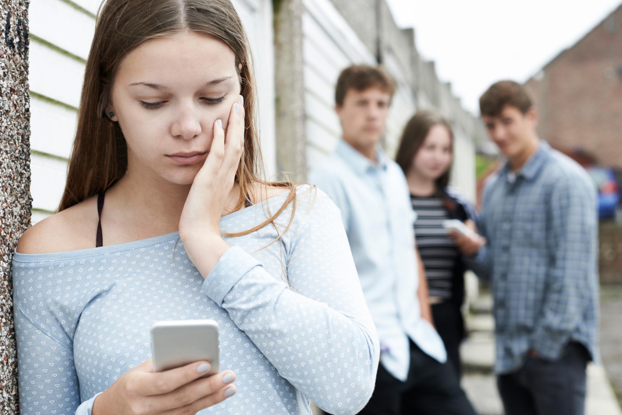 Junge Frau steht absteits einer Gruppe mit drei jungen Menschen und blickt auf ihr Handy. Es wirkt, als ob die Gruppe abfällig über die spricht.