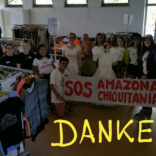 Das Bild zeigt einen Raum voller Kleidung. Es ist ein Markt für gebrauchte Kleidung. Im Raum stehen viele Menschen mit einer Fahne und einem Banner. Der Erlös geht in den Amazonas.