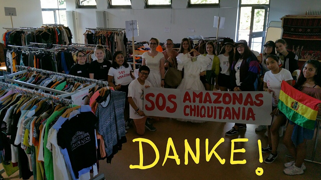 Das Bild zeigt einen Raum voller Kleidung. Es ist ein Markt für gebrauchte Kleidung. Im Raum stehen viele Menschen mit einer Fahne und einem Banner. Der Erlös geht in den Amazonas.