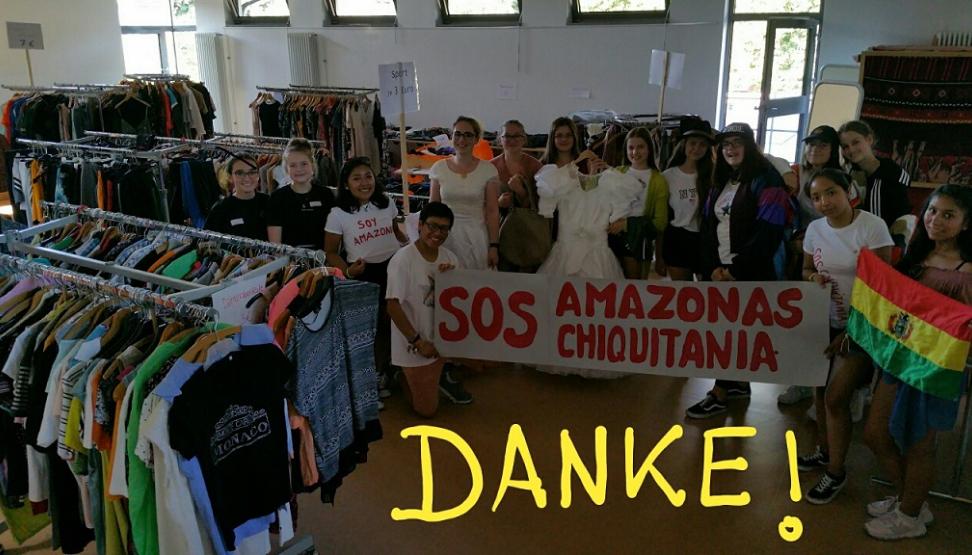 Das Bild zeigt einen Raum voller Kleidung. Es ist ein Markt für gebrauchte Kleidung. Im Raum stehen viele Menschen mit einer Fahne und einem Banner. Der Erlös geht in den Amazonas.