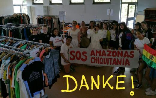 Das Bild zeigt einen Raum voller Kleidung. Es ist ein Markt für gebrauchte Kleidung. Im Raum stehen viele Menschen mit einer Fahne und einem Banner. Der Erlös geht in den Amazonas.