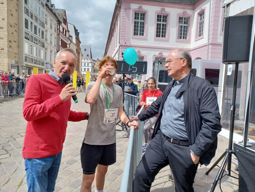 Das Bild zeigt den Bischof mit Moderator und Läufer während eines Spendenlaufes vor dem Trierer Dom. Man sieht viele Läufer.