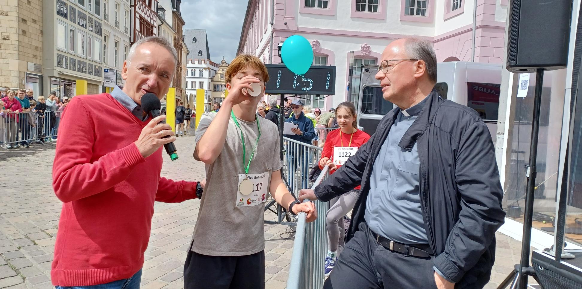Das Bild zeigt den Bischof mit Moderator und Läufer während eines Spendenlaufes vor dem Trierer Dom. Man sieht viele Läufer.