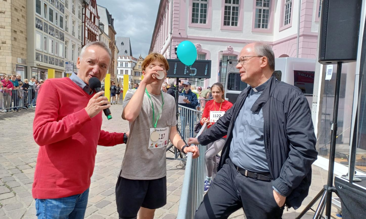 Das Bild zeigt den Bischof mit Moderator und Läufer während eines Spendenlaufes vor dem Trierer Dom. Man sieht viele Läufer.