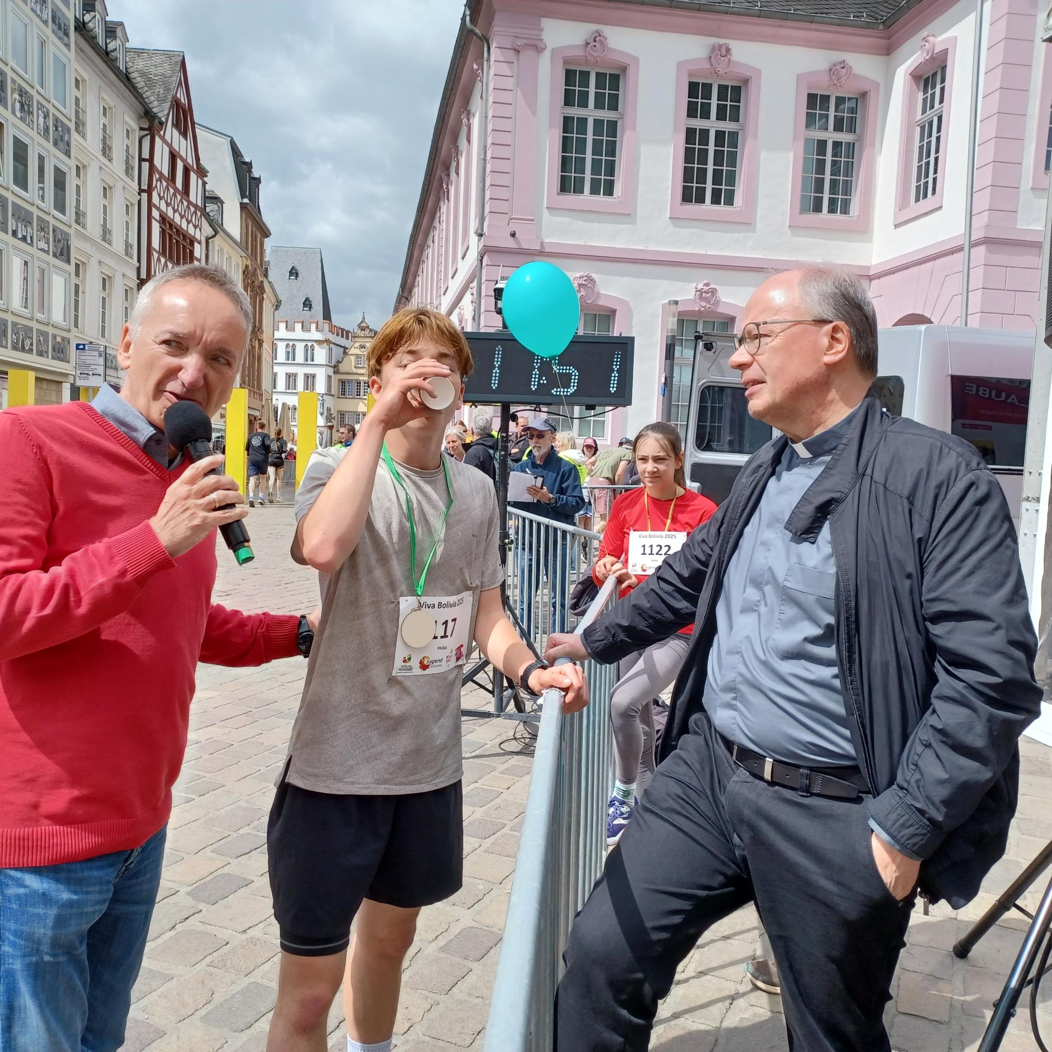 Das Bild zeigt den Bischof mit Moderator und Läufer während eines Spendenlaufes vor dem Trierer Dom. Man sieht viele Läufer.