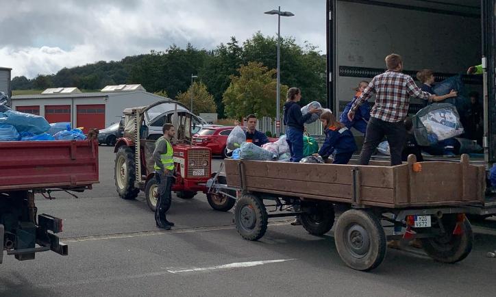 Das Bild zeigt einen Traktor mit Hänger vor einem LKW. Vom Hänger aus werden Säcke in den LKW geladen.