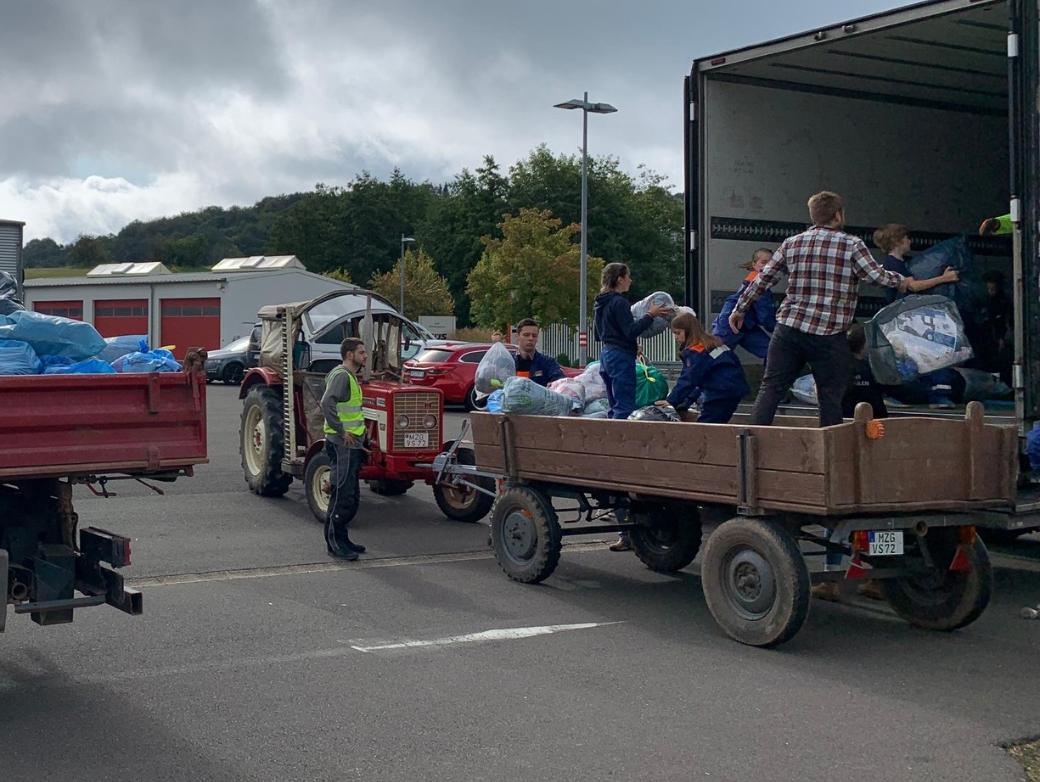 Das Bild zeigt einen Traktor mit Hänger vor einem LKW. Vom Hänger aus werden Säcke in den LKW geladen.