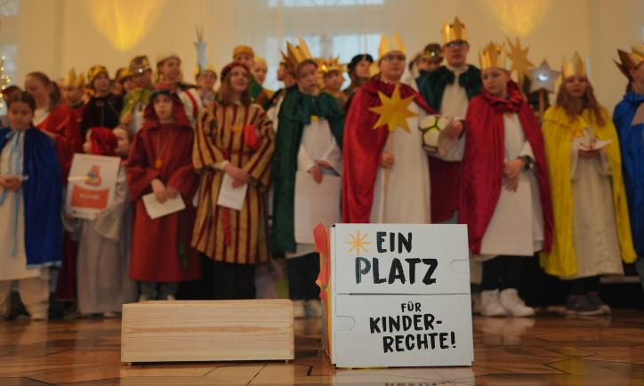 Das Bild zeigt eine Gruppe Sternsinger. Davor steht ein Schild mit der Aufschrift 'Ein Platz für Kinderrechte'