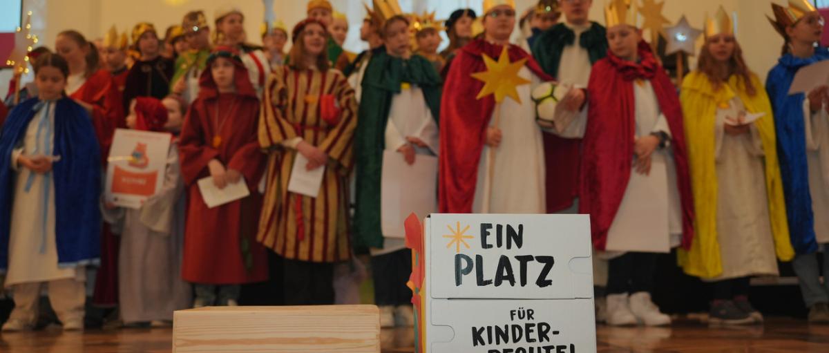 Das Bild zeigt eine Gruppe Sternsinger. Davor steht ein Schild mit der Aufschrift 'Ein Platz für Kinderrechte'
