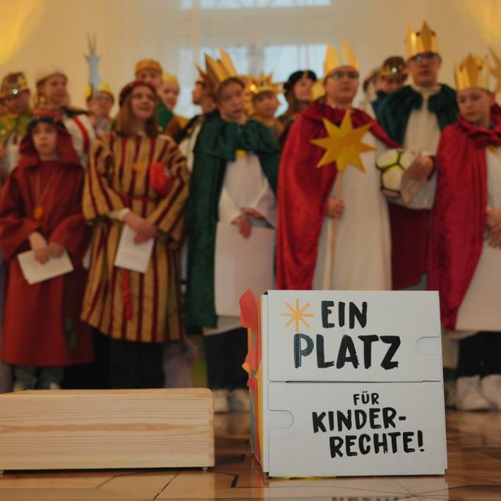 Das Bild zeigt eine Gruppe Sternsinger. Davor steht ein Schild mit der Aufschrift 'Ein Platz für Kinderrechte'