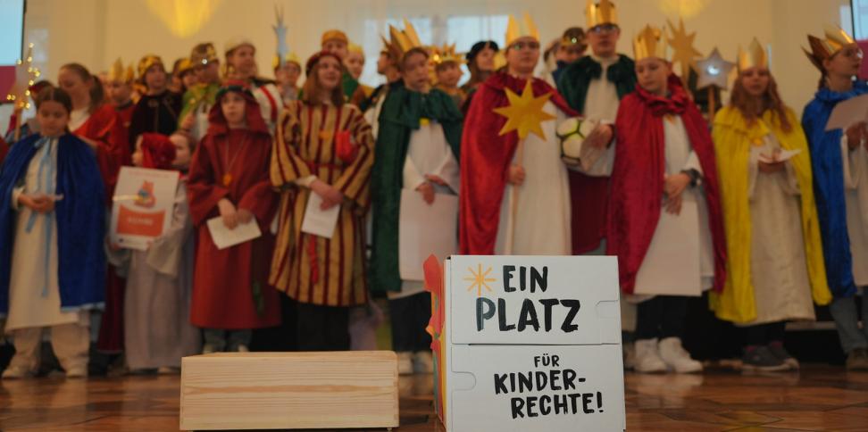 Das Bild zeigt eine Gruppe Sternsinger. Davor steht ein Schild mit der Aufschrift 'Ein Platz für Kinderrechte'