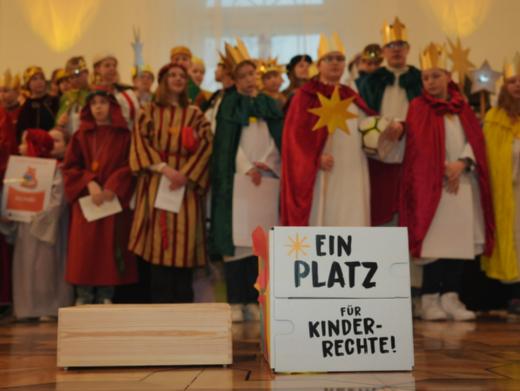 Das Bild zeigt eine Gruppe Sternsinger. Davor steht ein Schild mit der Aufschrift 'Ein Platz für Kinderrechte'