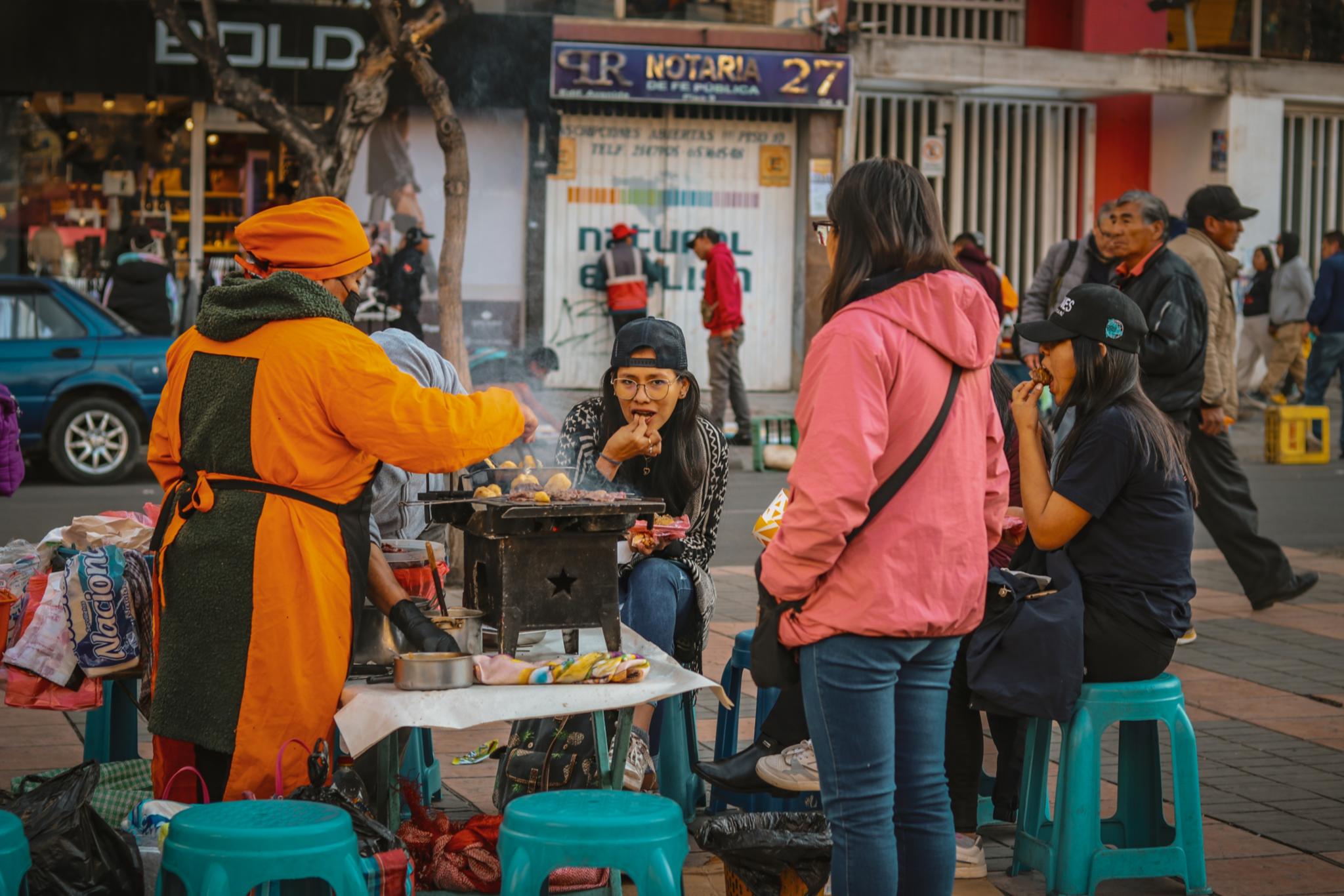 Das Bild zeigt einen Essensstand auf einer Straße in Bolivien. Darauf bereiten von hinten zu sehen zwei Personen zu und von vorne zu sehen ist eine Frau die isst.