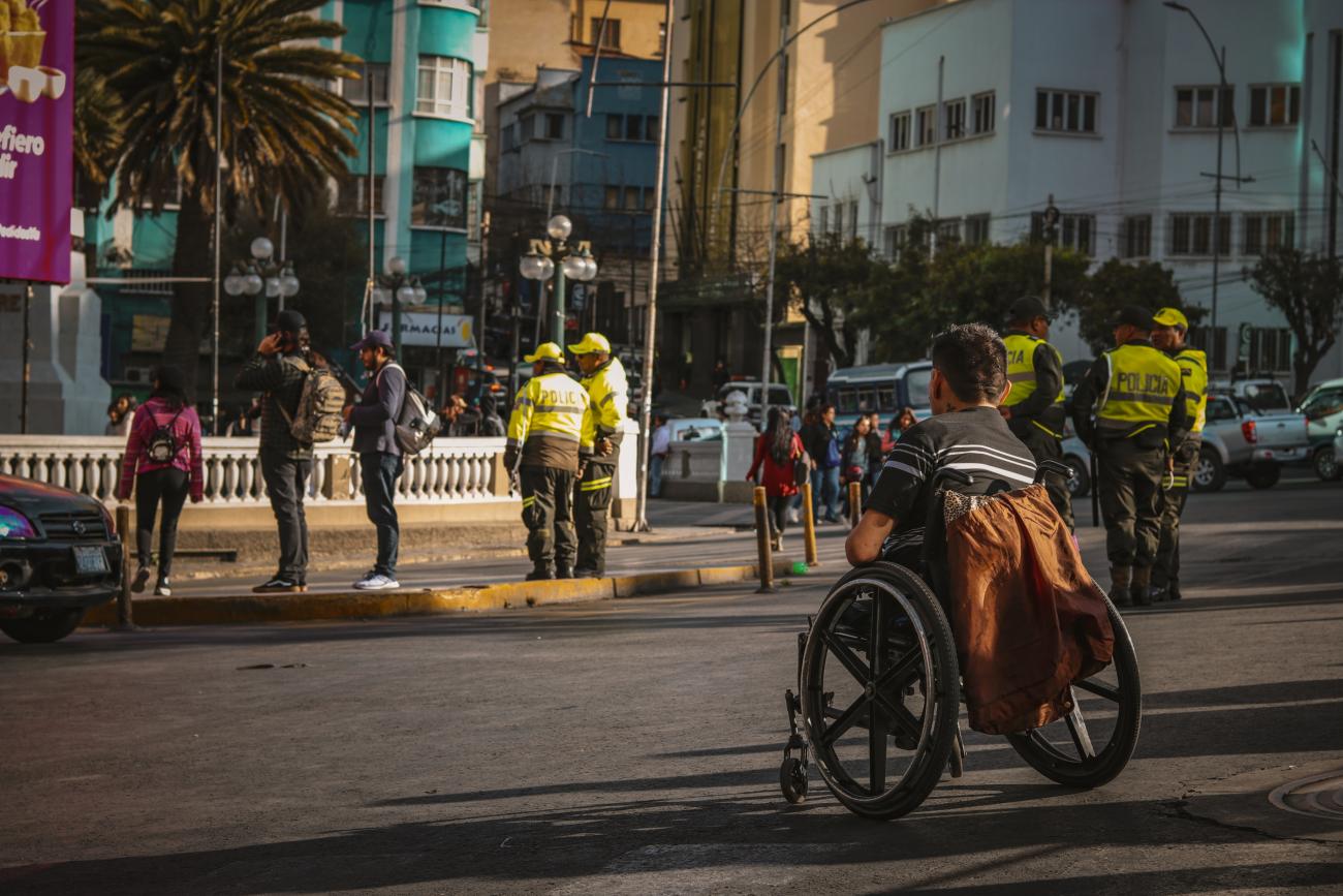 Das Bild zeigt einen jungend Mann im Rollstuhl sitzend mitten auf einer Straße vor einer Bordsteinkante.