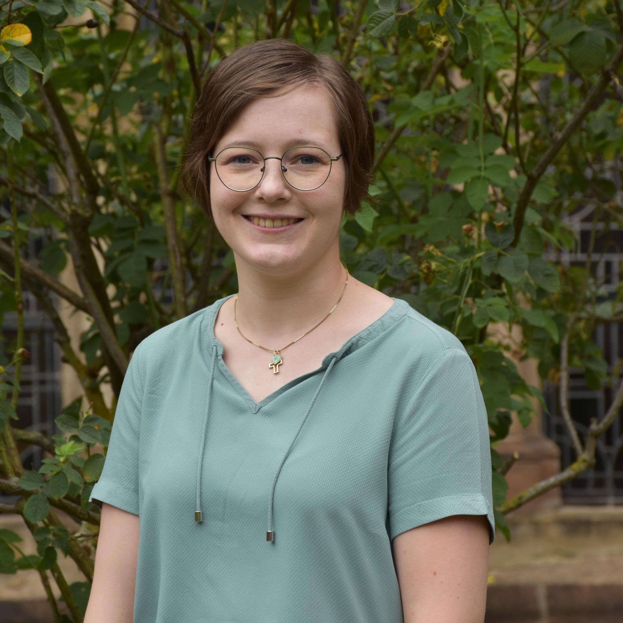 Das Bild zeigt ein hüfthohes Portrait einer jungen Frau mit Brille und braunen Haaren.