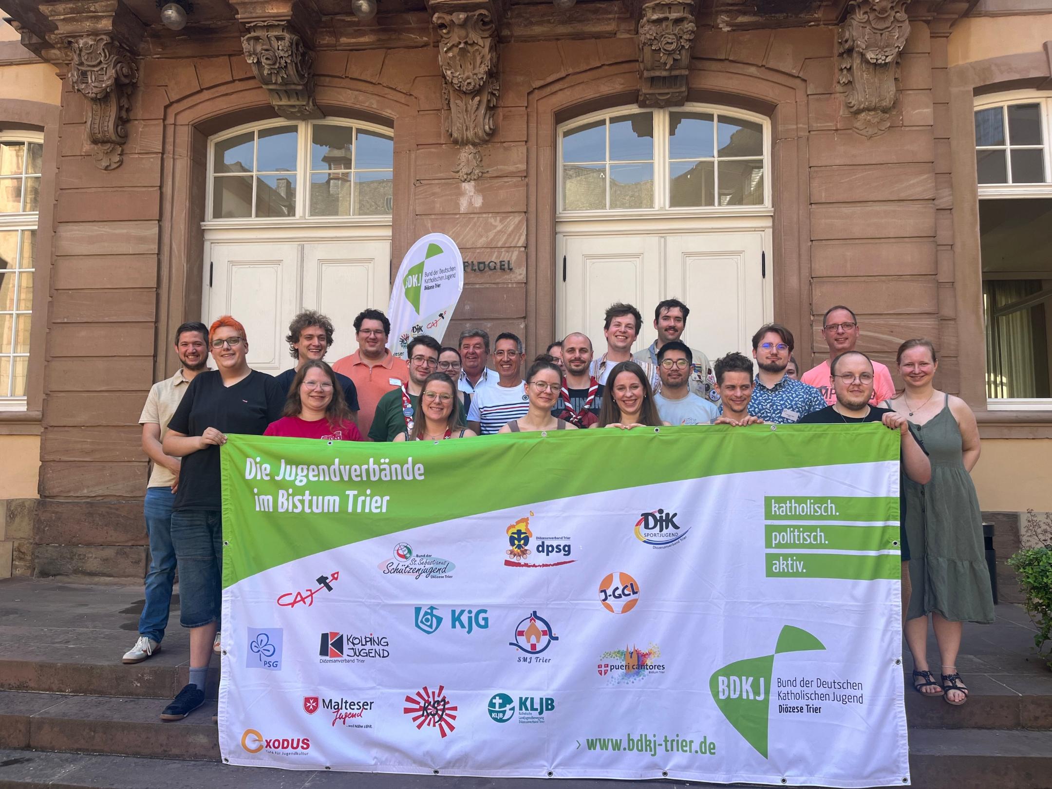 Das Foto zeigt ein Gruppenbild. Die Gruppe steht vor einem Gebäude und im Vordergrund wird eine Fahne hochgehalten. Auf dieser sind Logos von Jugendverbänden im Bistum Trier. Es ist das Gruppenbild zur Diözesanversammlung des BDKJ Trier.