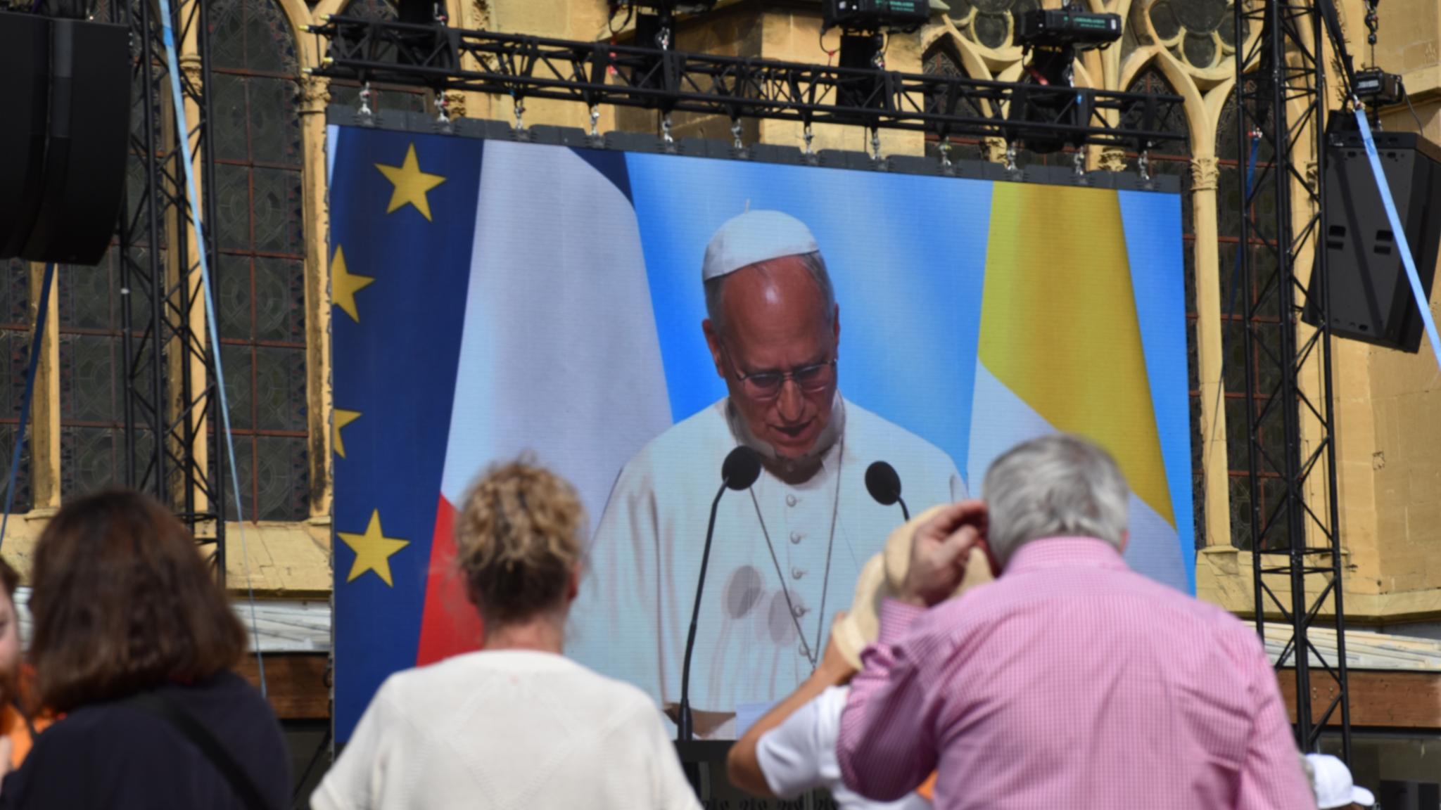 Viele Menschen stehen vor einem Großen Bildschirm. Er zeigt den Papst bei einer Rede vor verschiedenen Fahnen.